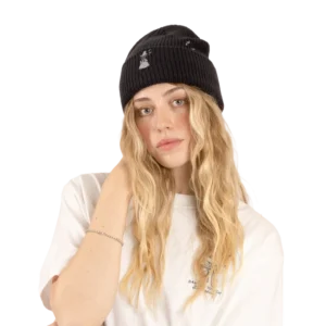 A woman with long, wavy blonde hair wears a grey Moomin beanie and a white T-shirt, accessorised with Moomin keyrings clipped to her bag. She is looking at the camera with a neutral expression, standing against a plain light grey background. Gate21.ee