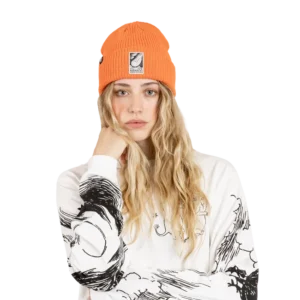 A person with long wavy blonde hair wears the Moomin Beanie in orange and a white sweatshirt with black graphic designs. Moomin keyrings dangle from the strap of their bag, adding a playful touch to their look. They have a neutral expression and rest their chin on their hand, looking straight at the camera. Gate21.ee