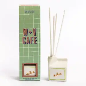 A box labelled W+V Cafe and Moon stands next to a white diffuser with four reeds. The diffuser, called Happy Cat - 100ml Diffuser from the Wick & Vessel home fragrance collection, has a green-bordered label with the word dolce written in yellow. Both items are set against a clean white background. Gate21.ee
