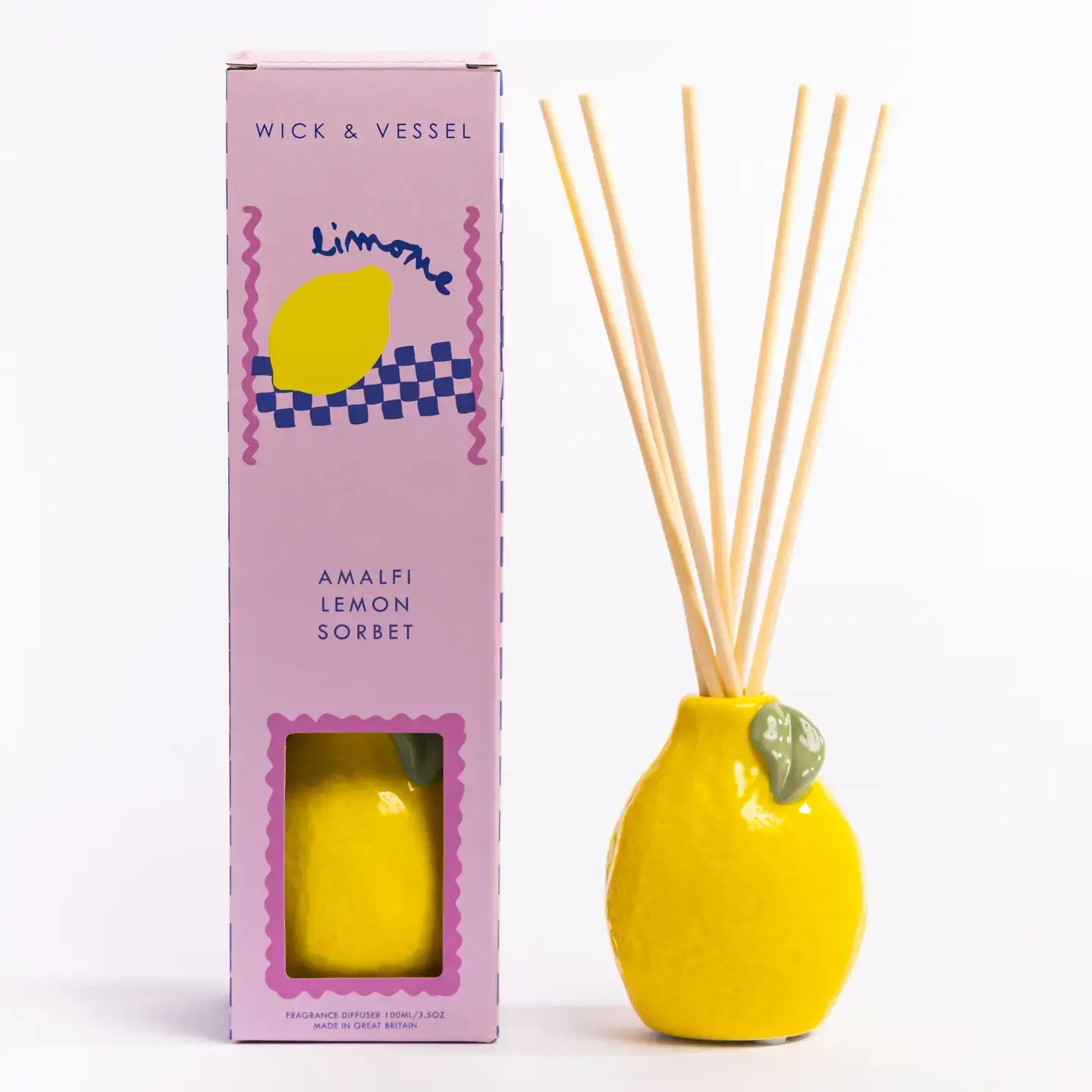 A lemon-shaped ceramic diffuser with reed sticks stands next to a pink and purple Happy Cat - 100ml Diffuser box labelled Wick & Vessel. This elegant Wick & Vessel home fragrance set features a charming box adorned with a lemon illustration and a window that reveals the beautifully crafted Happy Cat diffuser inside. Gate21.ee