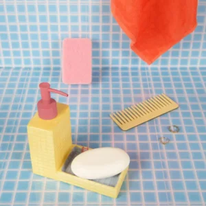 A yellow Pool - Soap Dispenser, a white bar of soap, a beige comb, two rings, a pink scourer pad, and an orange towel are artistically arranged on light blue tiled surfaces in a bathroom setting—an arrangement reminiscent of L’expressionist gifts that celebrate the beauty of everyday moments. Gate21.ee