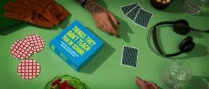A game box titled Things They Don’t Teach You At School sits on a green table surrounded by gloves, playing cards, headphones, and snacks. Gate21.ee